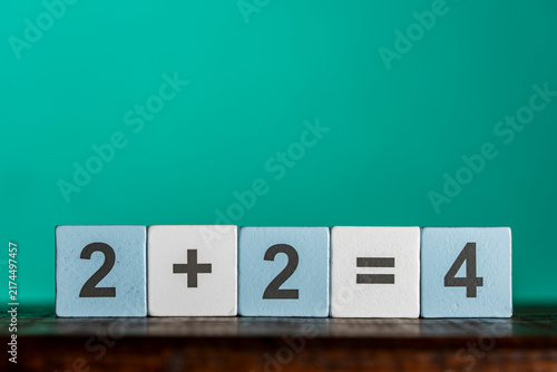 Simple math equation two plus two equals four on wooden toy blocks. Row of light blue and white wooden blocks spelling out basic arithmetic addition formula 2 plus 2 equals 4 on green background