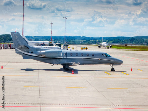 Business jet, painted in a silver-grey livery at the tarmac. A white airplane in the background. Midsize, twin-engine corporate jet. grey exterior paint scheme with rear-mounted turbofan engines. 