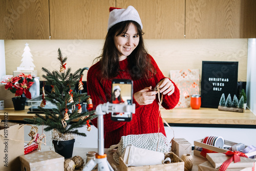 Foto Woman in Santa hat ties raffia ribbon before phone on tripod near CHRISTMAS VIBES ONLY sign