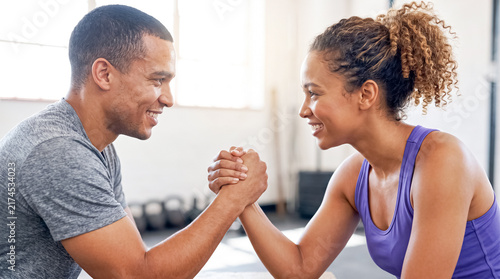 Friendly Arm Wrestling Match: A man and woman, both fit and smiling, engage in a friendly arm wrestling match, representing friendly competition and shared activity in a fitness setting.