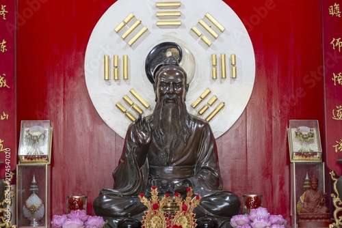 A dark, finely crafted statue of Laozi (Lao Tzu), the legendary philosopher and founder of Daoism, set against a large Yin-Yang symbol and surrounded by the eight Bagua symbols. Red plaques with golden characters and offerings, Chee Chin Khor Temple, Bangkok, Thailand