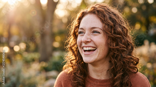 Joyful young woman with red curly hair and freckles laughing outdoors in warm sunlight