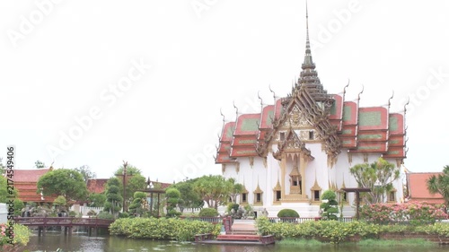 traditional thai architectural structure beside a tranquil canal with a red pedestrian bridge, greenery and landscaped surroundings in thailand