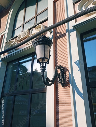 Canvastavla Vintage black wrought iron street lantern mounted on a classic building facade with large glass windows and decorative architectural elements under bright sunlight