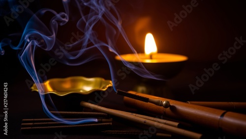 Close up of burning incense sticks releasing swirling blue smoke near glowing candlelight with aroma