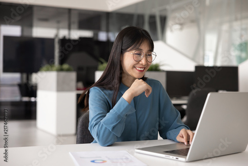 Foto Smiling young Chinese professional woman, office employee using laptop sit at de