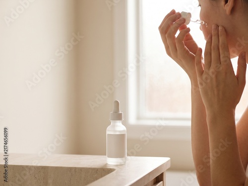 Minimalist skincare routine with face serum bottle, woman applying facial oil in bright bathroom, natural beauty wellness concept