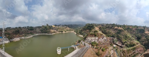 Fotografía High-angle aerial panoramic view of a water reservoir and dam surrounded by green hills, infrastructure buildings, and a dramatic cloudy sky during the day