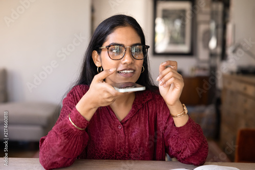 Fotografía Young Indian woman sitting at table at home, speaking into her smartphone using voice assistant, recording audio message or leaves voicemail