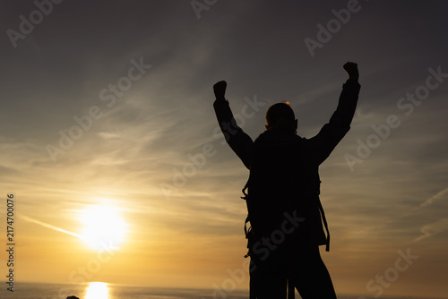Obraz na płótnie Hiker silhouette standing on a cliff at sunset, raising arms in celebration of a