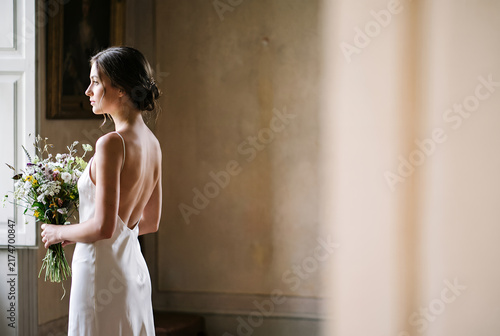 Young woman in white dress holding bouquet by window