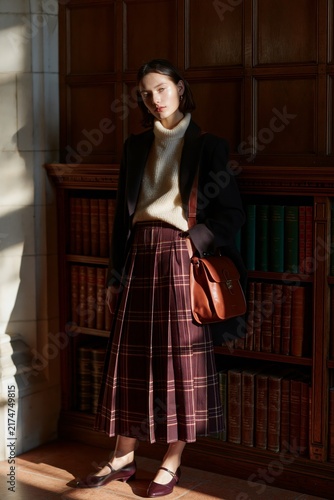 Fashion editorial featuring a woman in a dark academia outfit with a plaid skirt and sweater, standing beside a wooden bookshelf filled with books