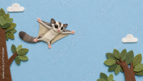 A cute handcrafted felt sugar glider soars between leafy trees beneath soft white clouds in a blue sky.