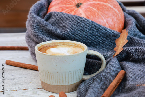 Cup of spicy coffee, orange pumpkin wrapped in gray warm scarf, cinnamon sticks, dry fall leaves close up. Autumn hot drink, November morning concept