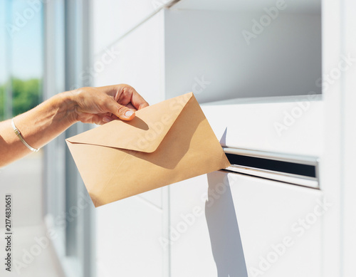 A hand inserts a brown envelope into the slot of a white mailbox. This close-up view captures a person sending mail or a document, showing postal service action on a sunny day with a modern exterior.
