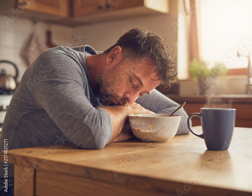 A tired, exhausted man sleeping at the kitchen table during breakfast with a bowl of cereal and coffee mug, feeling sleepy from burnout and early morning fatigue in a cozy home interior.