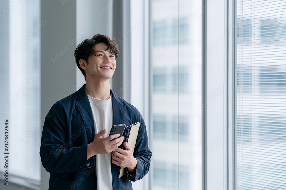 Fototapeta premium 笑顔で窓の外を眺めるスマートフォンを持つ若手ビジネスマン
