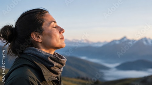 A woman wearing a neck gaiter and jacket admires a misty mountain valley landscape during the soft light of golden hour