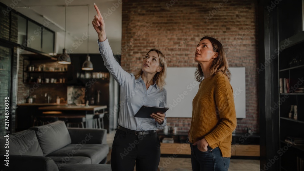 Obraz premium A female realtor in a blue shirt points upward while holding a tablet, guiding a client in a mustard sweater through a loft with brick walls and hanging pendant lights