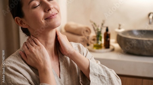 A young woman in a robe gently massages her neck and shoulders in a bright bathroom with standing minimalist bathroom