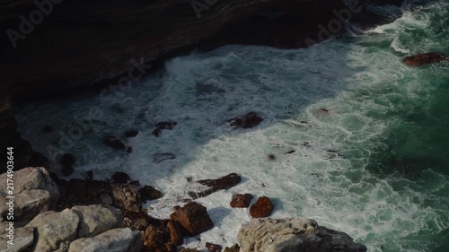 Turquoise ocean waves surge and break over dark coastal rocks, creating frothy white patterns and vivid green water in a rugged shoreline scene, with natural motion and textured seascape detail.
