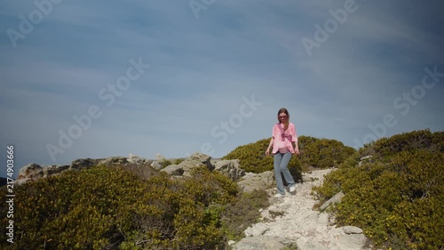 Young woman hikes down a rocky coastal trail, wearing casual city clothes as she walks between low green shrubs under a blue sky, blending urban style with an active solo outdoor escape.