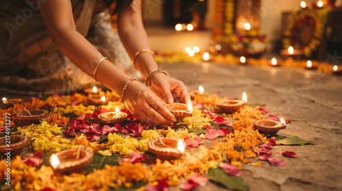 Woman in traditional attire arranging lit diyas on colorful flower rangoli pattern during deepavali festival celebration at home.