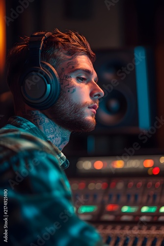 Tattooed man wearing headphones in a music recording studio looking at a mixing console board