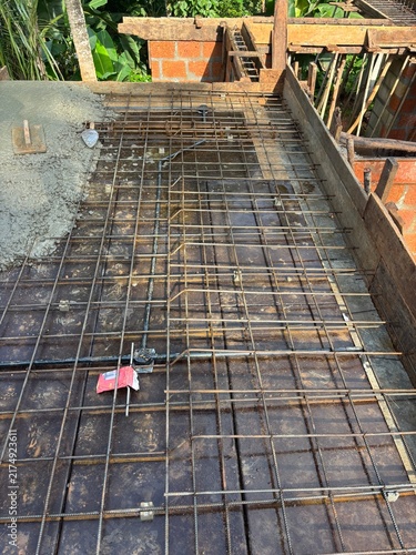 House construction site in Kerala, India, showing a partially built residential home with concrete columns, exposed brick walls, construction materials, and workers’ activity.