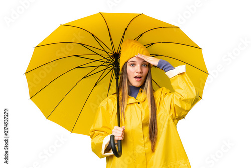 Teenager girl with rainproof coat and umbrella over isolated chroma key background doing surprise gesture while looking to the side
