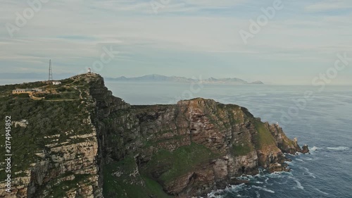 Drone orbit around Cape Point shows rugged sandstone cliffs, green coastal vegetation, foaming Atlantic waves, lighthouse and distant mountains beneath softly clouded sky, highlighting dramatic se##