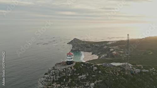 Aerial drone view of Cape Point lighthouse above rocky Atlantic coast, turquoise coves, winding trails, ocean cliffs, and warm low sunlight creating tranquil South African seascape at sunset.
