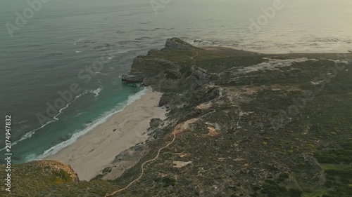 Drone view reveals wild coastline with rocky headland, pale sandy beach, turquoise shallows, winding trails and low vegetation, while white surf meets open water in warm hazy light.