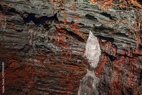 Foto A clear icicle hangs against folded red and black lava inside Raufarholshellir near the South Coast, Iceland