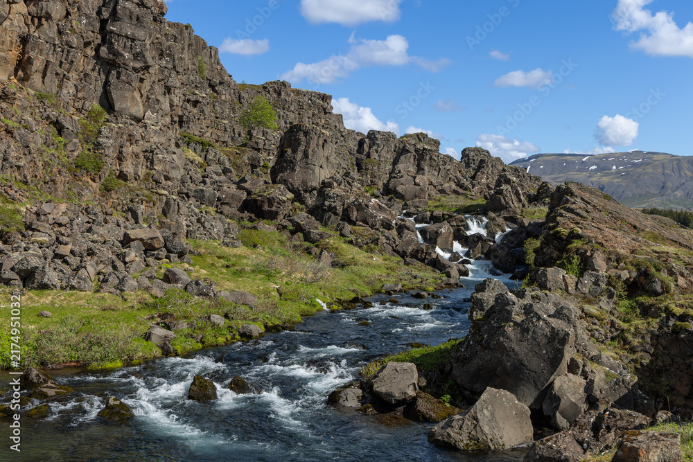 Fototapeta premium Several shallow drops through the rift channel appear within Thingvellir National Park along the Oxara River in Iceland. Water steps diagonally across basalt ledges beneath scattered clouds.