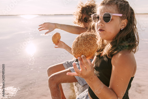 children holding fresh bread rolls while sitting on a driftwood log at shallow salt lake during warm summer sunset with relaxed family picnic atmosphere concept of family travel, bakery, tourism
