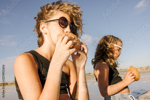 girls holding fresh bread rolls while sitting on a driftwood log at shallow salt lake during warm summer sunset with relaxed family picnic atmosphere concept of family travel, bakery, tourism