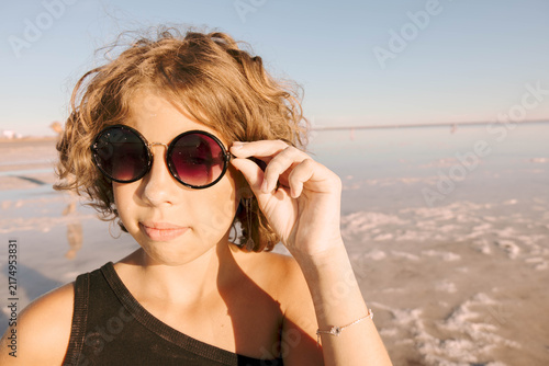 child adjusting sunglasses at pink salt lake, close up outdoor portrait with calm shoreline and natural summer landscape, travel lifestyle eyewear fashion tourism business