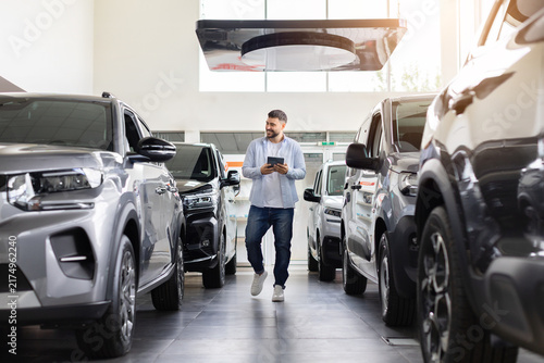 Cuadro en lienzo Young male customer walking through modern car dealership with tablet, comparing