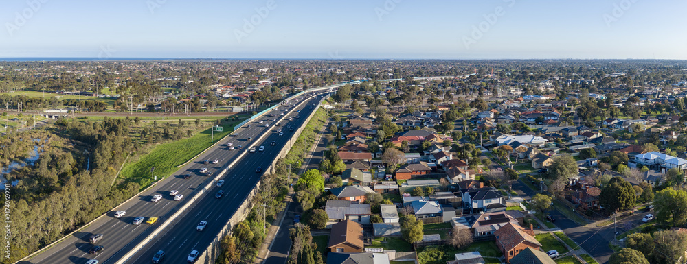 Obraz premium Aerial&nbsp;panorama view of Princes Freeway traffic bordering suburban residential housing, green reserves, and a railway line in Laverton, Melbourne, Australia. Traffic parallel to rail infrastructure