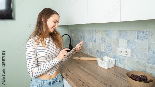 attractive smiling woman in kitchen at home relaxed using smartphone wearing white shirt and jeans