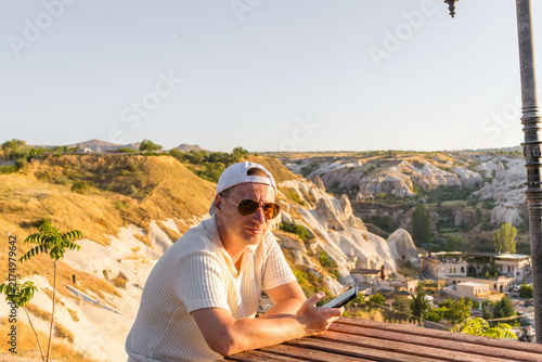Man with phone sitting at wooden table overlooking canyon. Digital travel represents lifestyle connection.