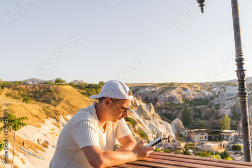 Man with phone sitting at wooden table overlooking canyon. Digital travel represents lifestyle connection.