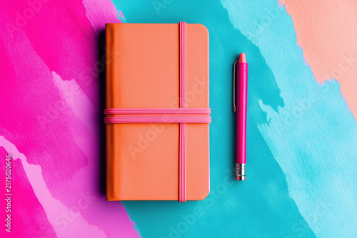 Orange leather notebook with elastic band and pink pen on a vibrant pink and teal background.