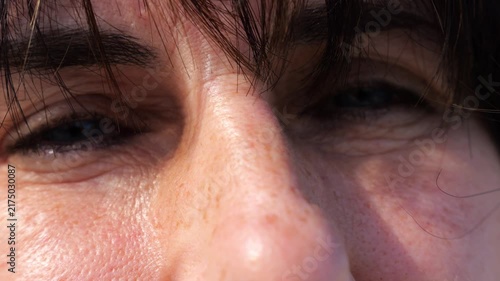 Tired blue eyes of a female blinking and looking into camera outdoor. Face of adult woman illuminating by bright rays of sun. Upset facial expression of lady. Close up Slow motion