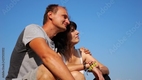 Happy pair hugs during sitting on wooden log on beach. Romantic couple admiring sea landscape at sunny day. Lovers spending time together at resort. Concept of summer vacation or holiday. Slow mo