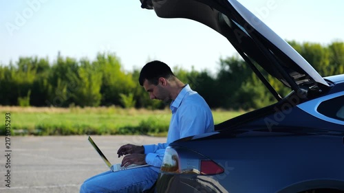 Young handsome entrepreneur works on notebook sitting in electric car trunk. Successful businessman in formal wear using computer at auto. Confident manager typing text on laptop outdoor. Slow mo
