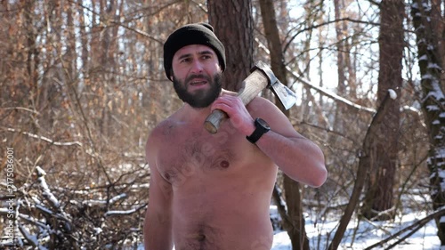 Portrait of shirtless bearded man stands in a snowy winter forest with axe. Tired guy resting after chopping logs in snowy woodland. The scene represents outdoor work in cold conditions. Close up