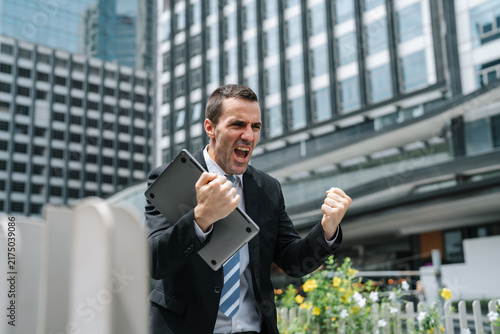 Businessman celebrating success with arms raised holding laptop outdoors, excited professional shouting in joy near modern office building, career achievement concept