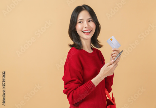 Happy Asian woman in a red sweater holding a credit card and smartphone while smiling, digital payment and festive shopping concept.